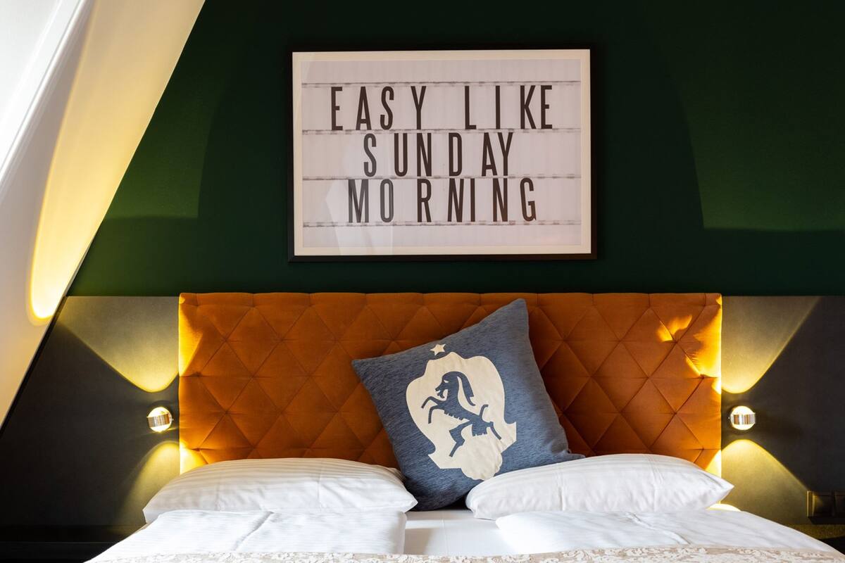 A neatly made bed with a decorative pillow, a framed sign above, and a warm-toned headboard.