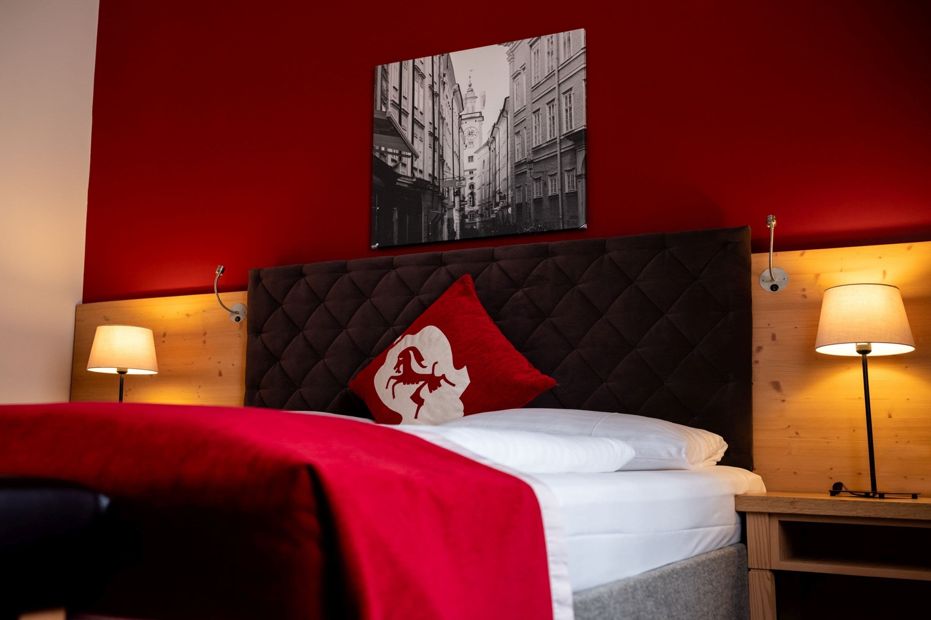 Ein Hotelzimmer mit roter Wand, einem Bett mit weißer Bettwäsche, einem rot-weißen Kopfkissen, zwei Nachttischlampen und einem schwarzen und weißen Foto im Rahmen über dem Bett.