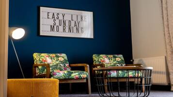Una habitación con una pared azul oscuro, un cartel enmarcado que dice "EASY LIKE SUNDAY MORNING", dos sillones florales, un reposapiés amarillo y una mesa redonda con sillas negras de alambre.