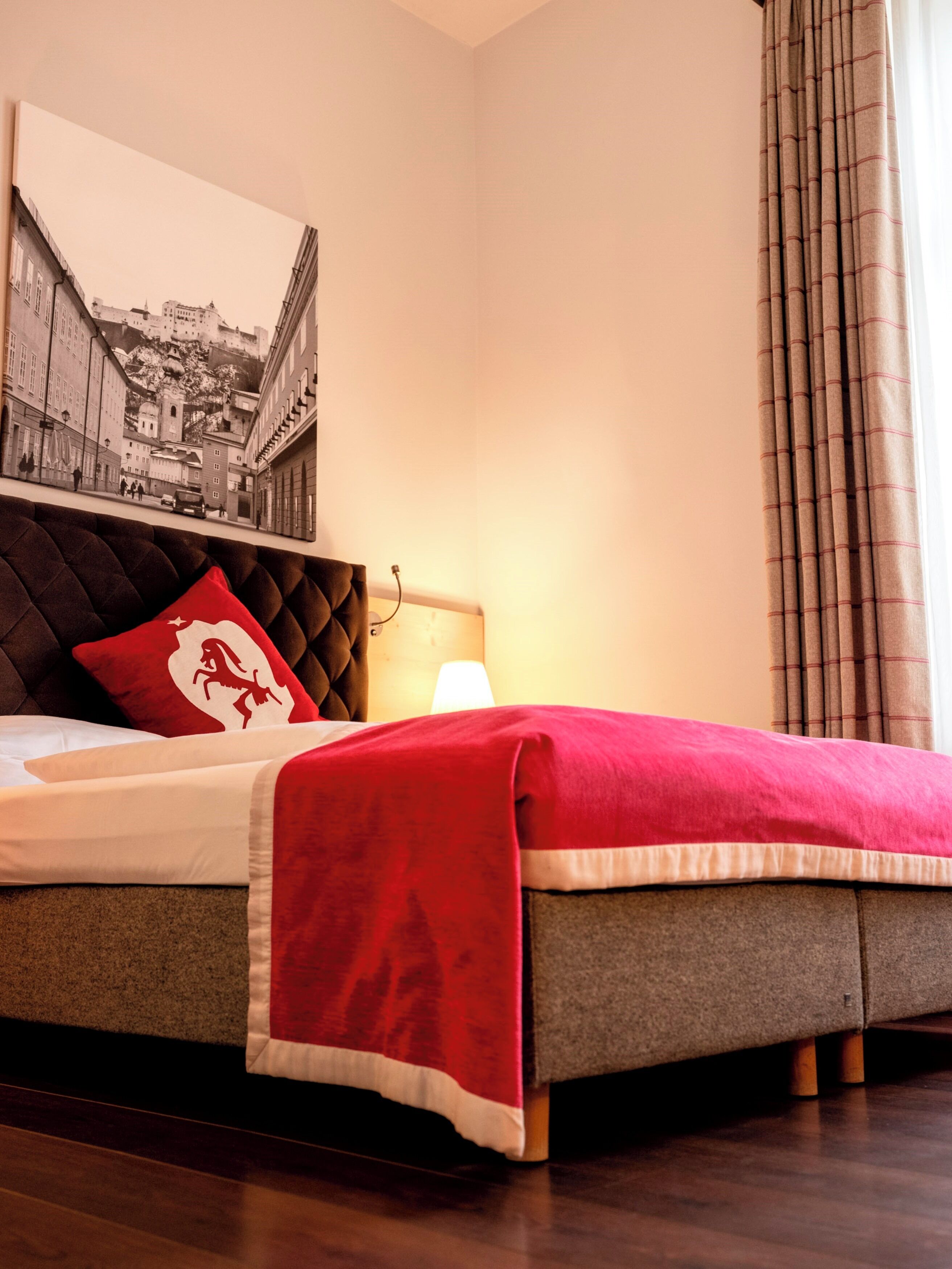 Un lit bien fait, recouvert d’une couverture rouge et agrémenté d’un oreiller aux motifs rouges et blancs. Une lampe est posée sur la table de chevet, et un tableau encadré est accroché au mur au-dessus du lit.