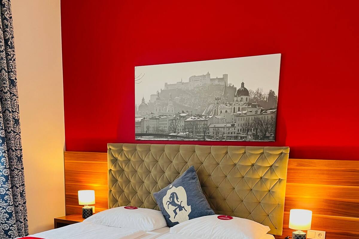 A hotel room with a large bed, two bedside tables with lamps, a red accent wall, and a cityscape painting above the bed.