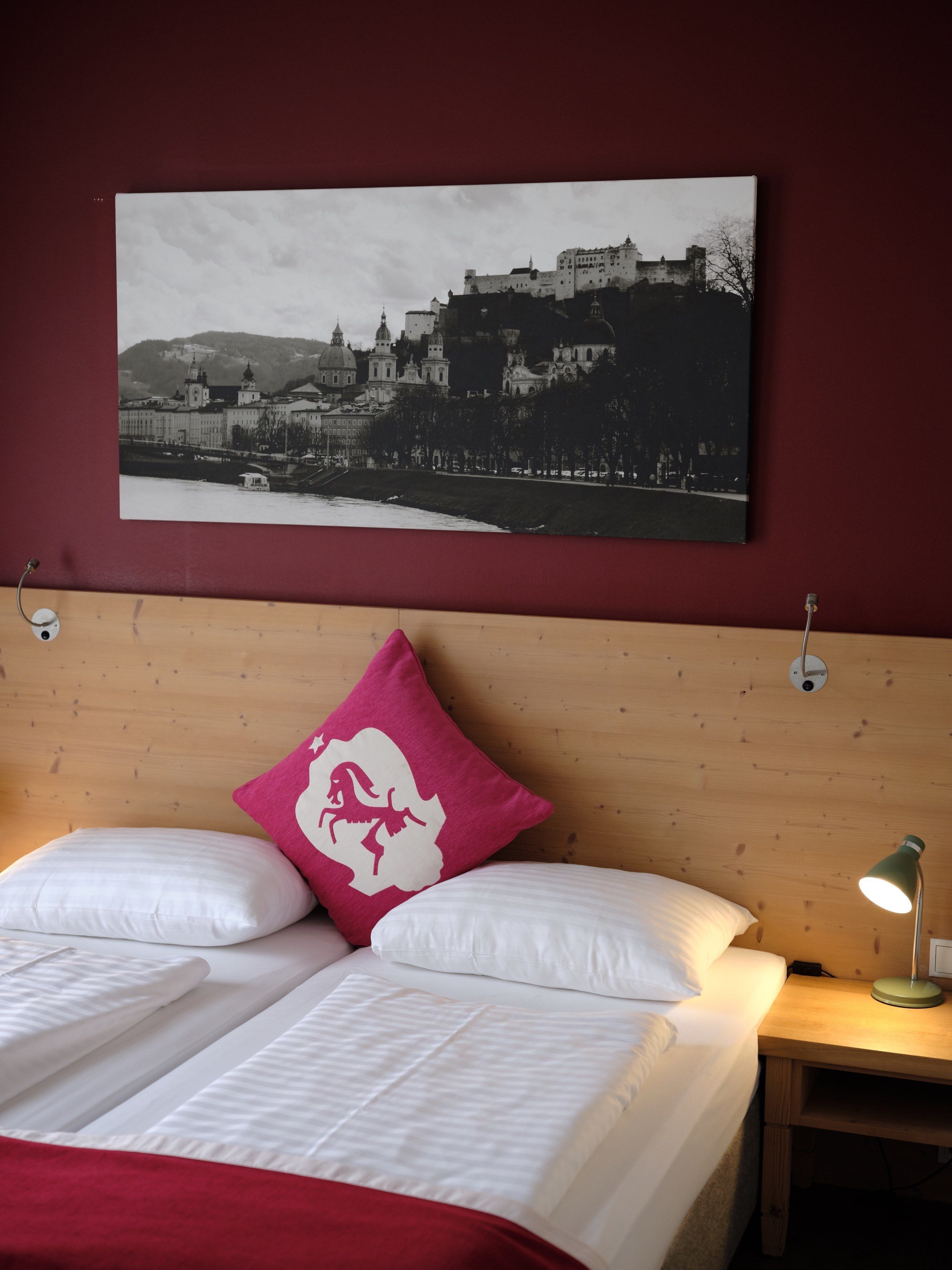 Un lit bien fait, avec du linge de lit blanc et un seul oreiller rose, une tête de lit en bois et un tableau représentant un bâtiment historique accroché au mur.