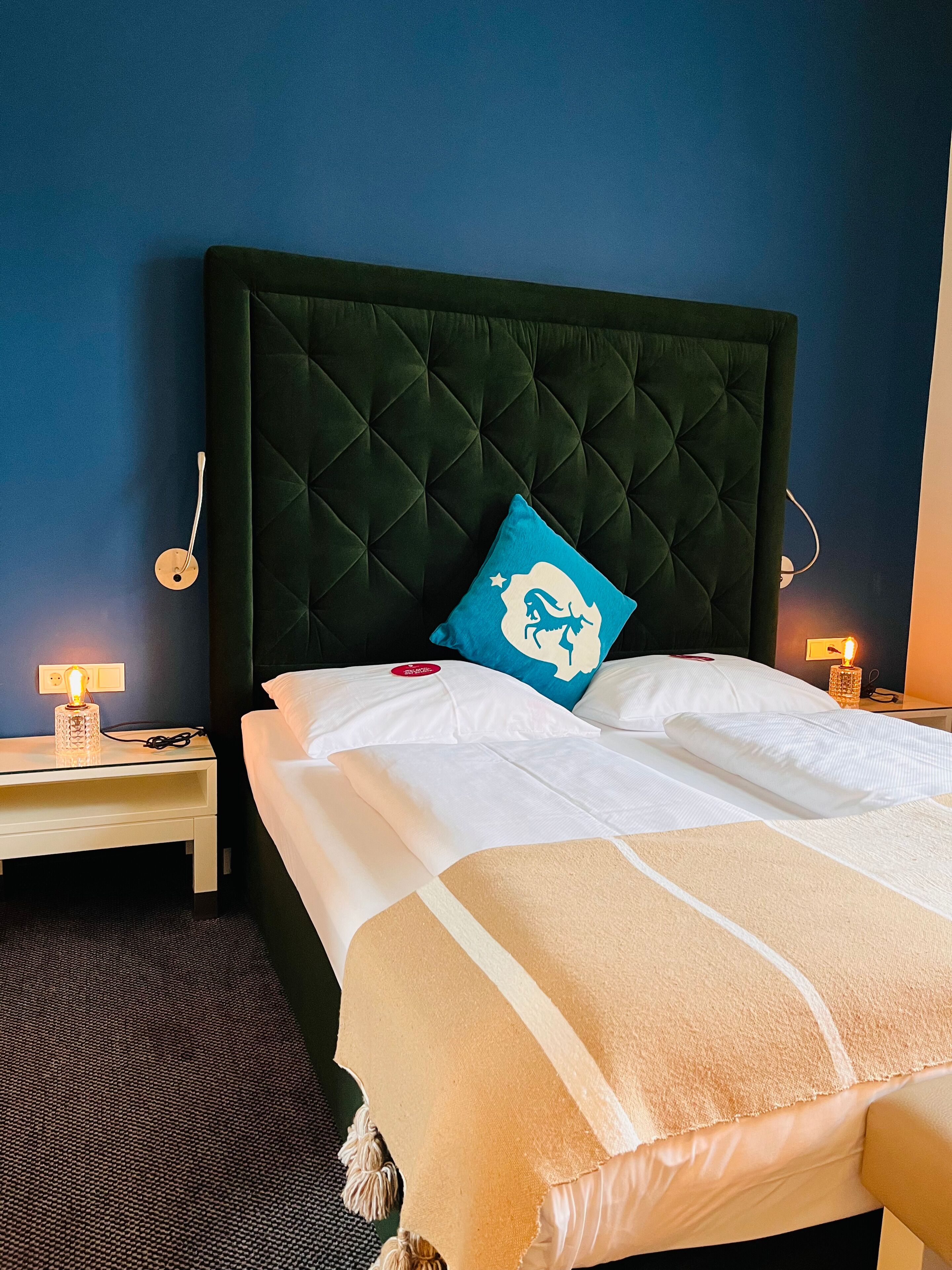 A neatly made bed with a dark headboard, white linens, and a blue pillow with a dinosaur design. There are two bedside lamps on either side of the bed.