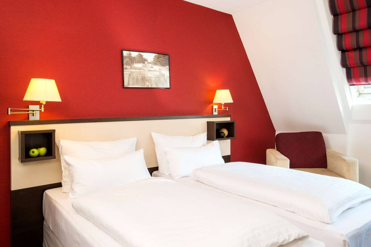 A hotel room with two beds, a red accent wall, two bedside lamps, a small shelf with apples, and a chair with a red cushion.