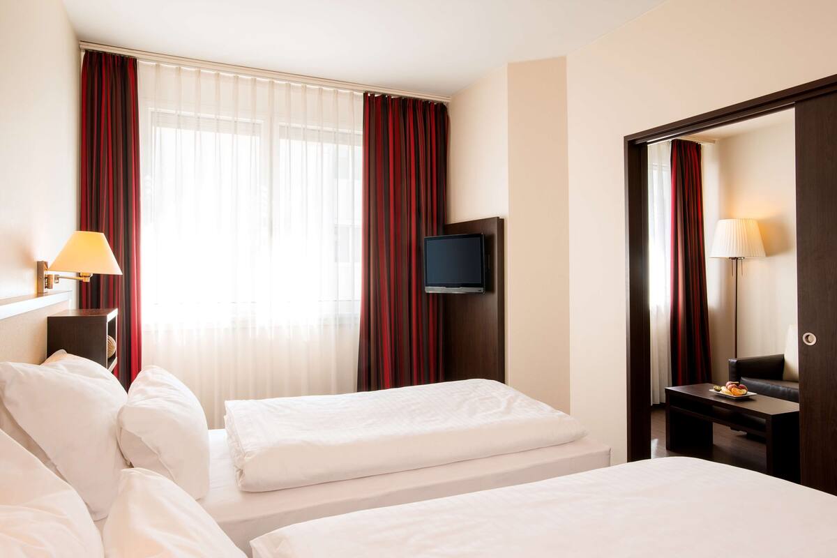 A hotel room with two beds, a TV, a desk with a lamp, and a view of the outside through sheer curtains.