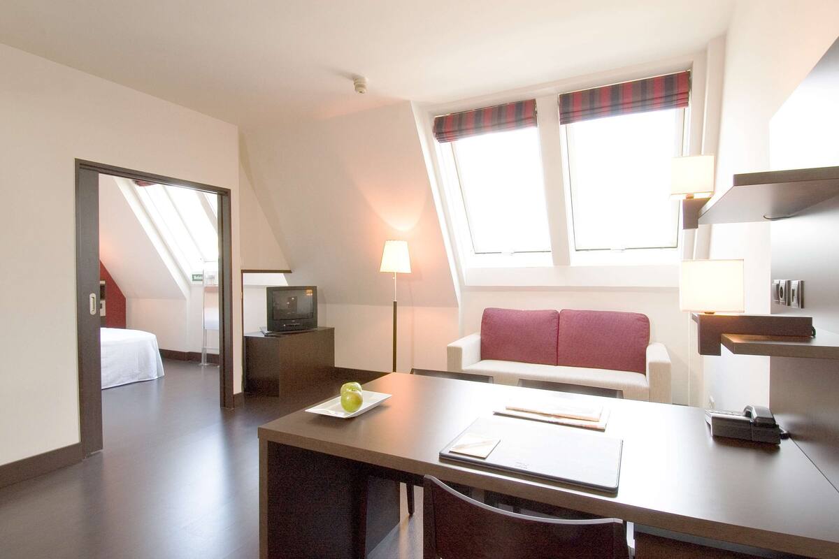 A hotel room with a dining area, a TV, a lamp, a window with a plaid curtain, and a bed in the background.