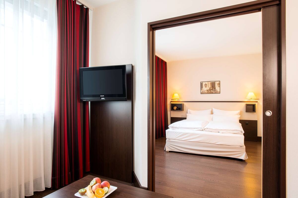 A hotel room with a bed, a television, a table with fruit, and red curtains.
