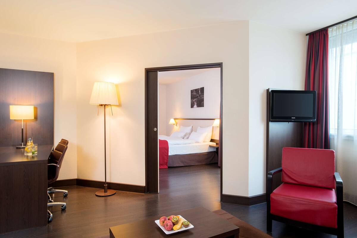 A hotel room with a desk, a chair, a bed, a television, and a dining table with fruit.