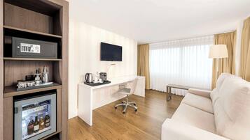 A hotel room with a wooden cabinet, a minibar, a desk with a chair, a television, a sofa, and a window with curtains.