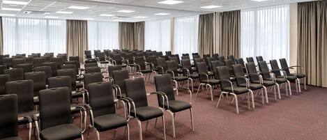 Un salón de conferencias con filas de sillas negras, amplias ventanas con cortinas y un suelo alfombrado en color rojo.