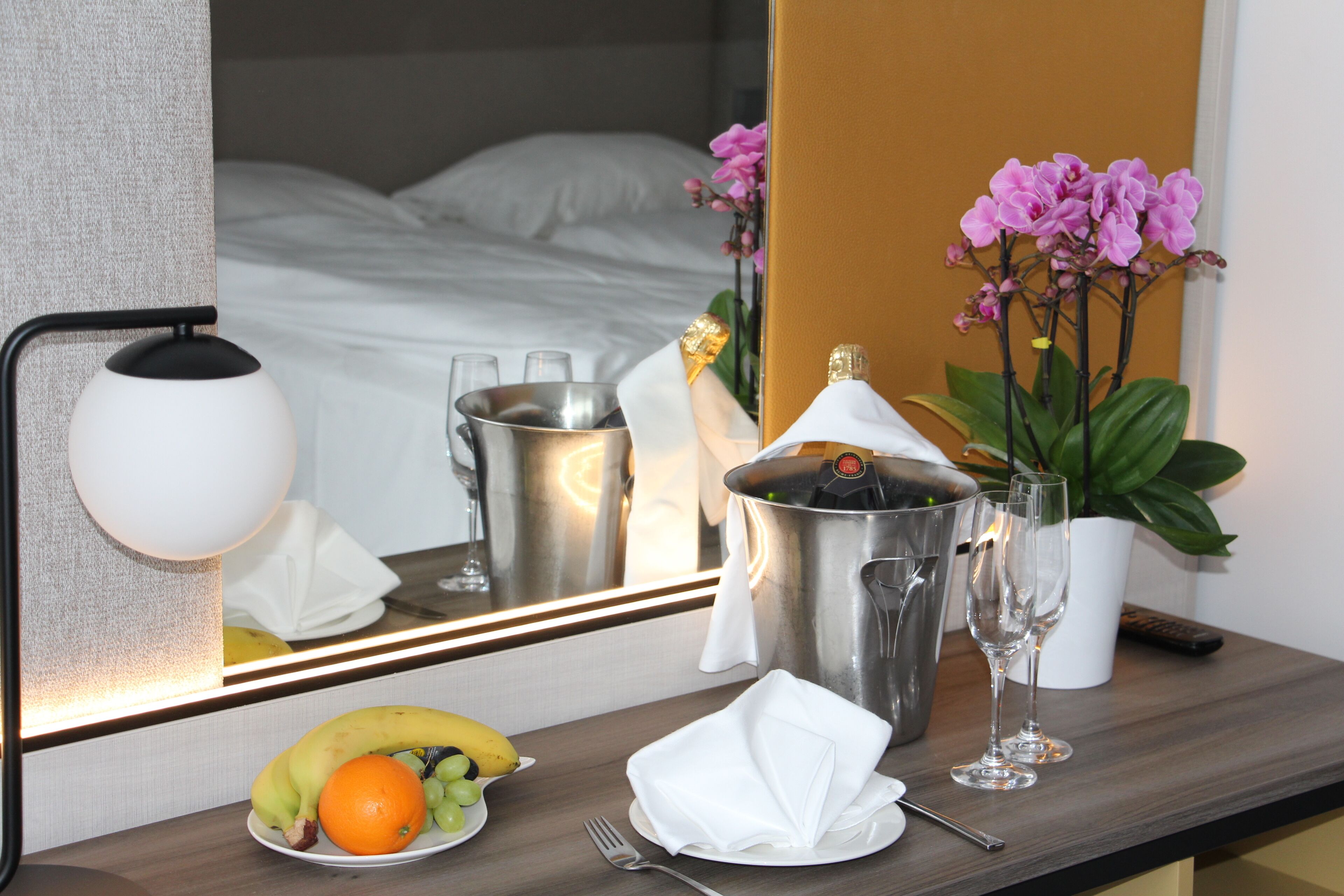 A hotel room with a bed, a bedside table with a lamp, a fruit bowl, a bottle of champagne, and a potted orchid.