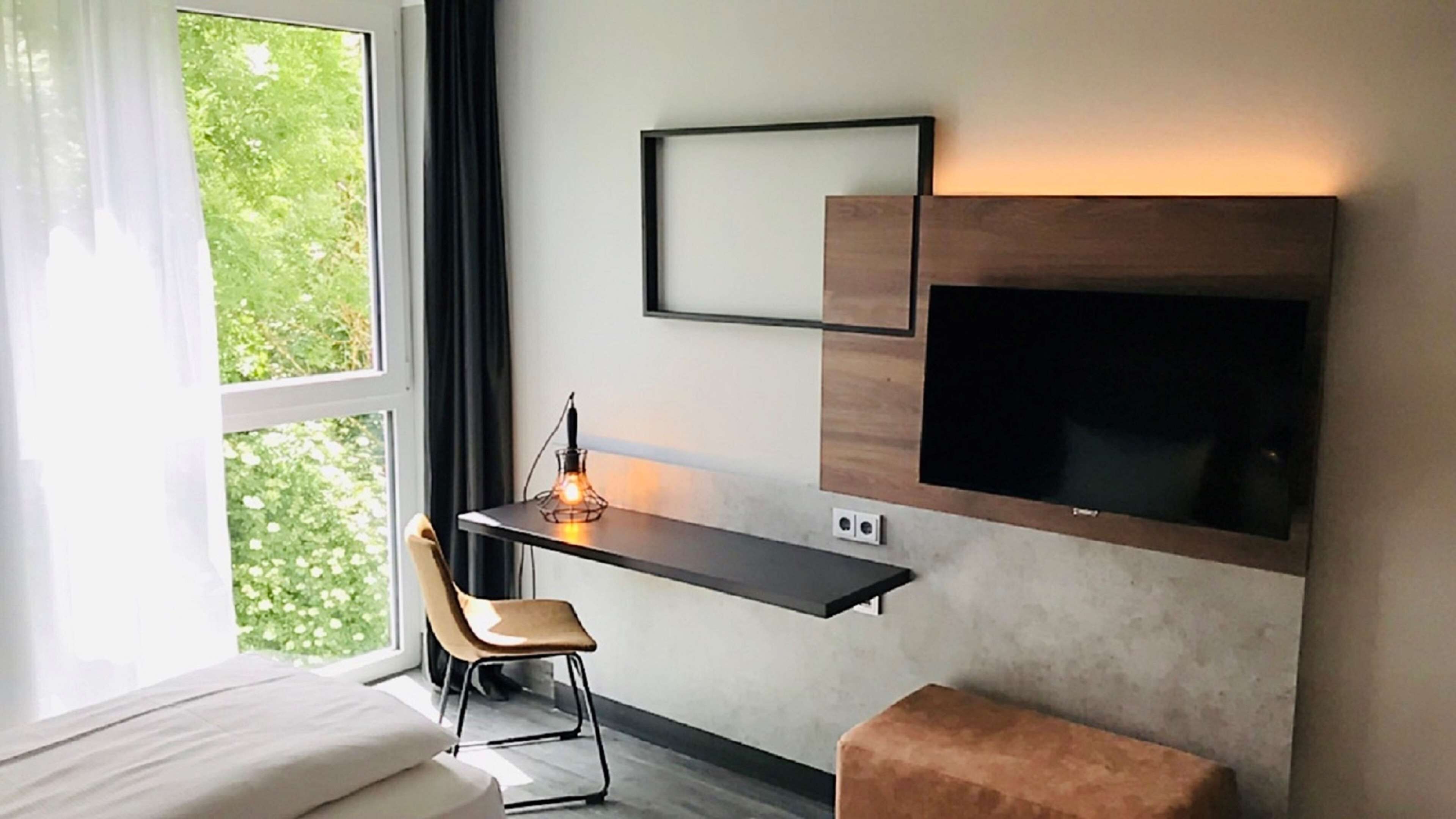 A modern hotel room with a bed, a desk with a chair, a TV mounted on the wall, and a window with a view of greenery.