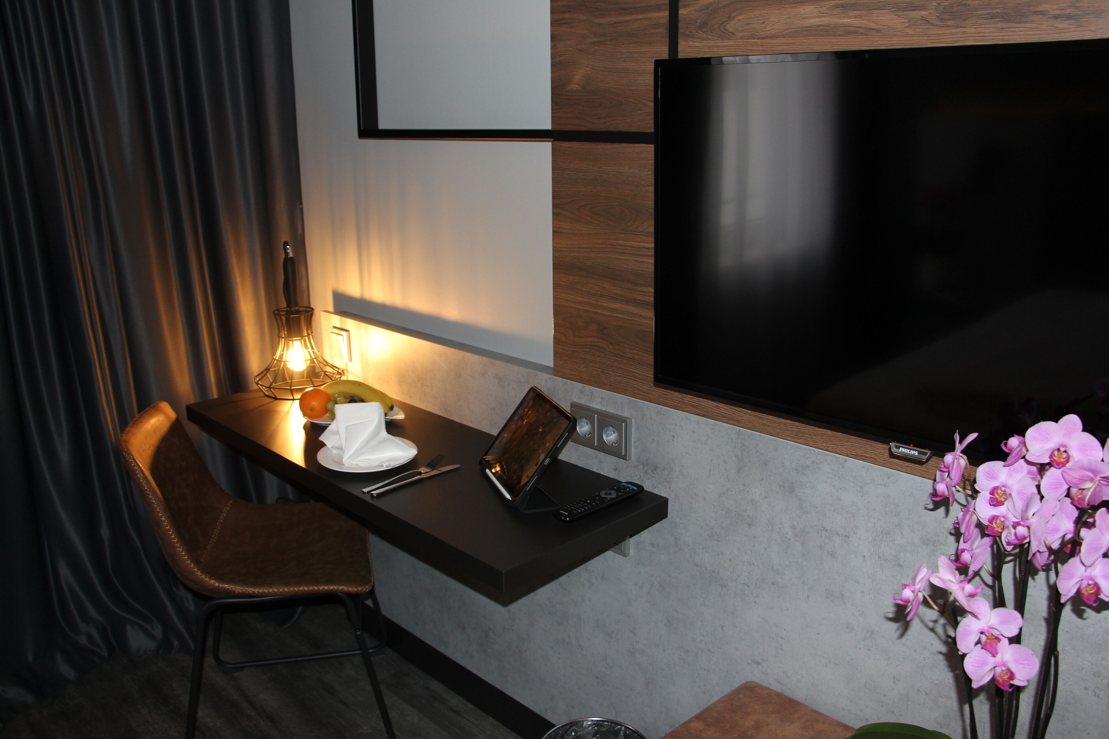 A modern hotel room with a flat-screen TV mounted on the wall, a small desk with a laptop, a chair, and a vase of pink orchids.