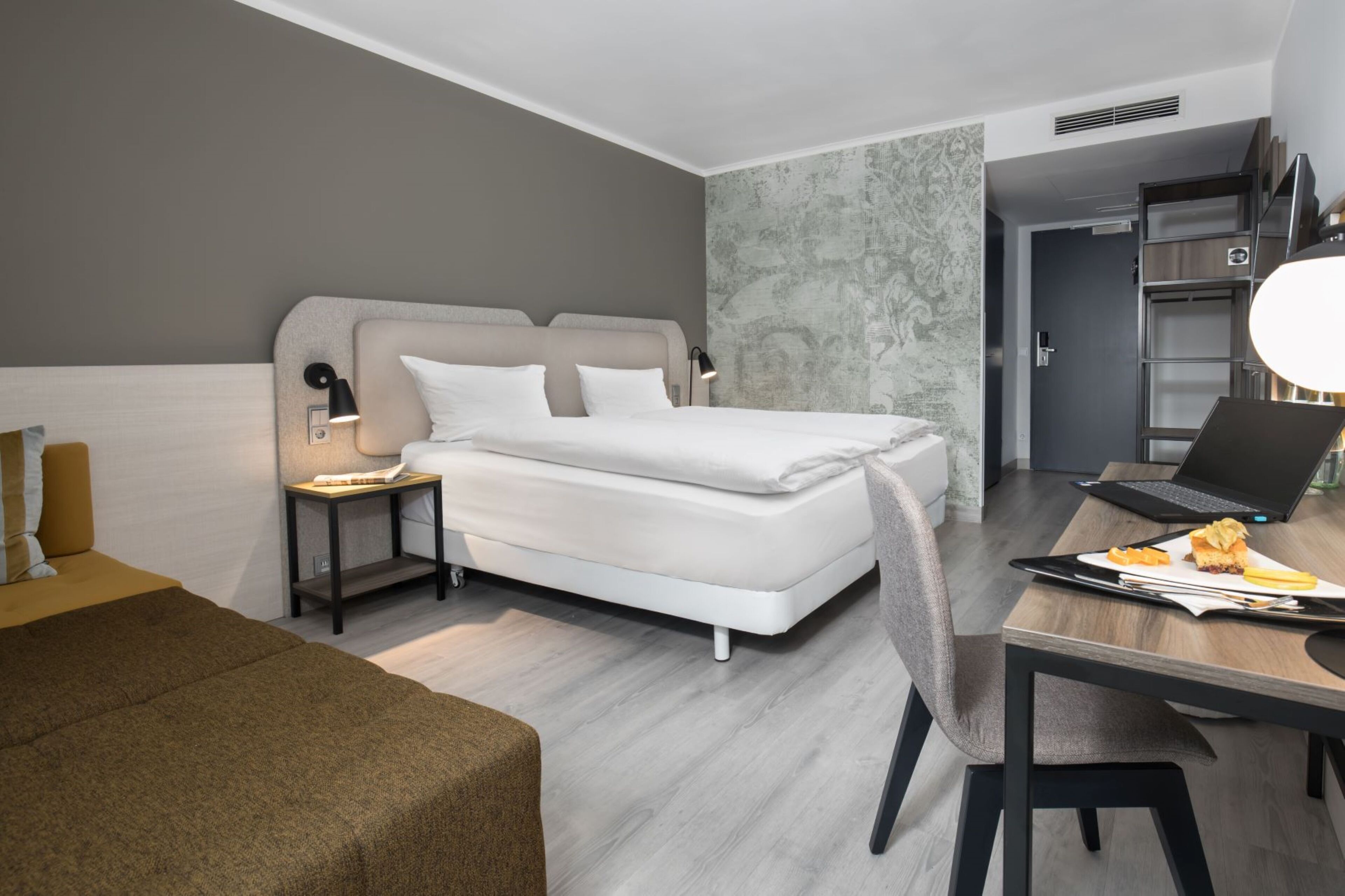 A modern hotel room with a bed, bedside table, lamp, laptop, and breakfast tray.