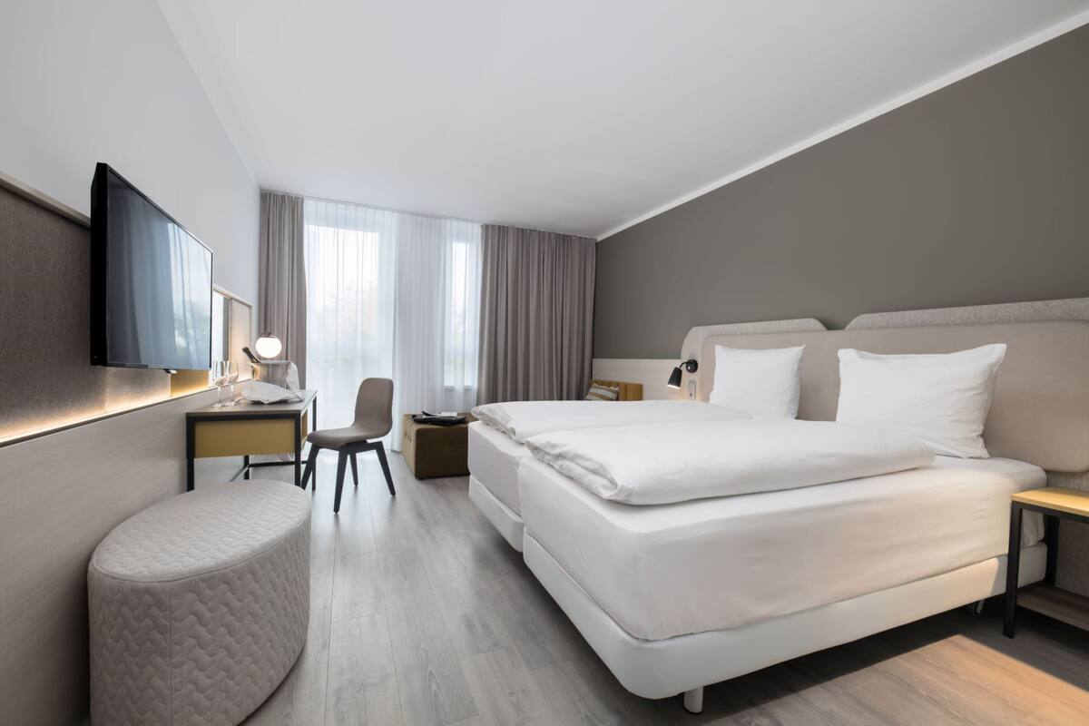 A modern hotel room with a large bed, a desk with a lamp, a chair, and a small ottoman.