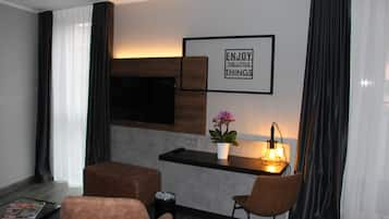 Una habitación de hotel moderna con una televisión de pantalla plana montada en la pared, una mesita con una lámpara, dos sillas, una planta en maceta y una frase motivacional en la pared.