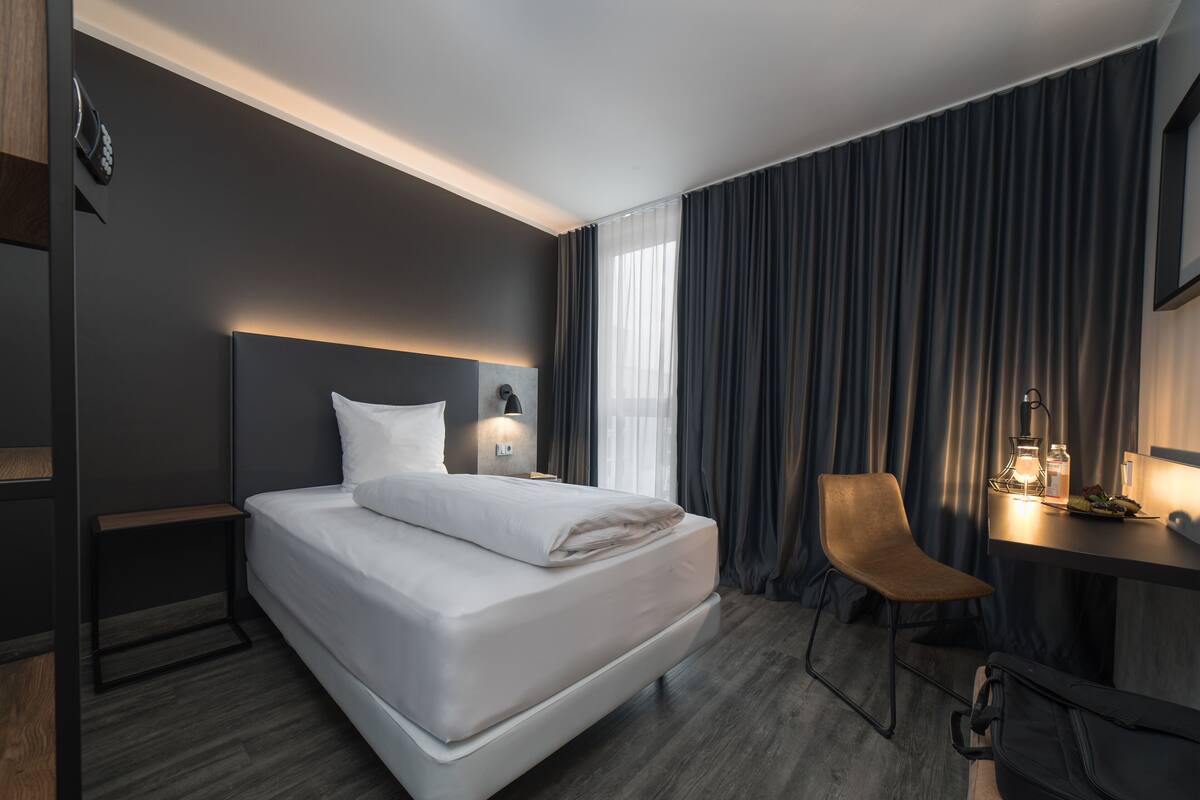 A modern hotel room with a large bed, a desk with a lamp, a chair, and a minibar.