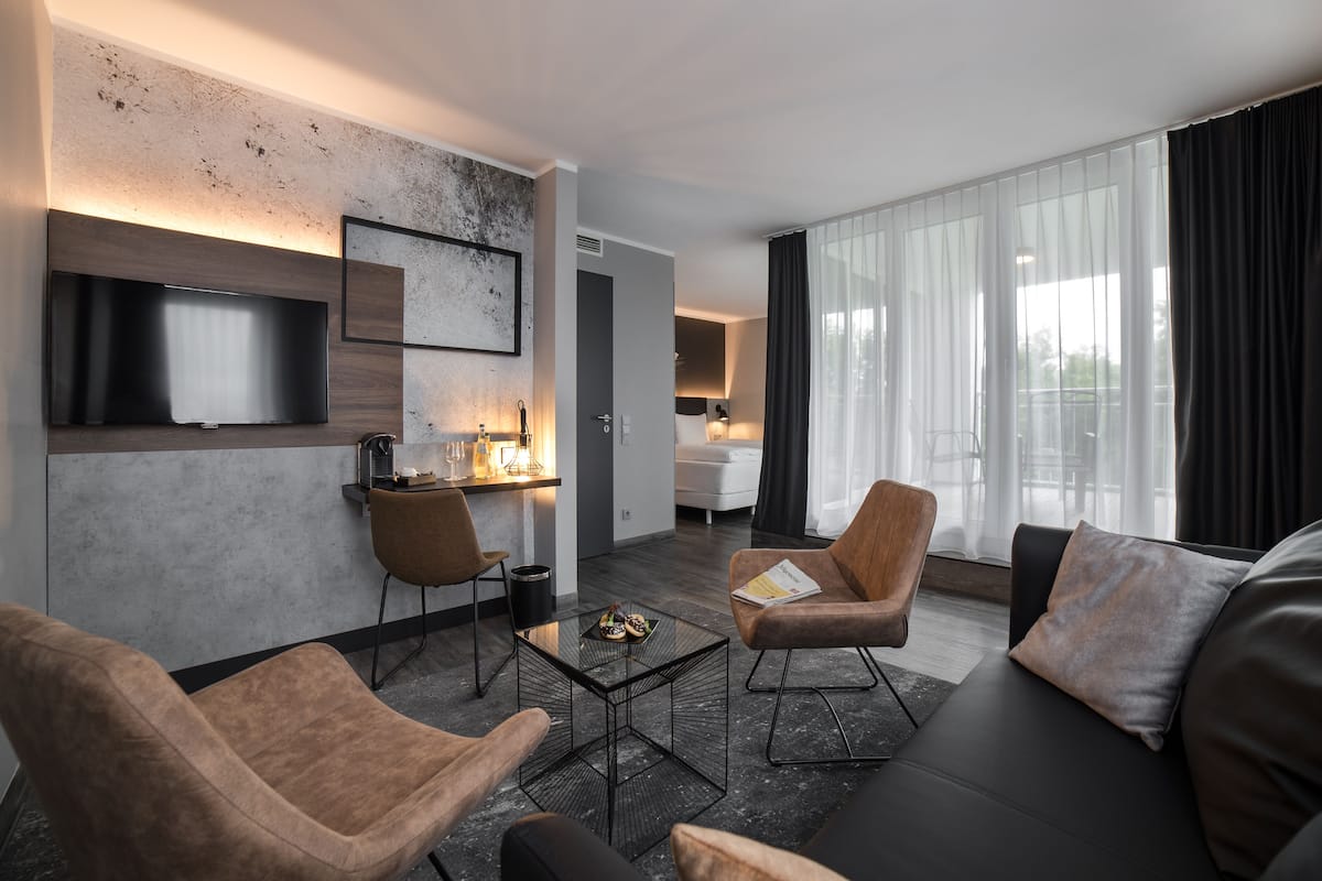 A modern hotel room with a living area featuring a sofa, two armchairs, a coffee table, and a flat-screen TV mounted on the wall.