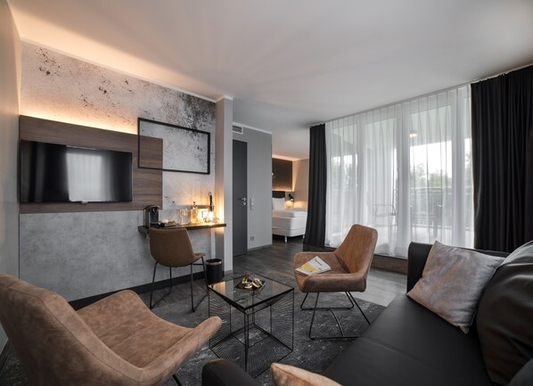 A modern hotel room with a living area featuring a sofa, two armchairs, a coffee table, and a flat-screen TV mounted on the wall. - SCOTTY + PAUL Hotel Deggendorf (Deggendorf)