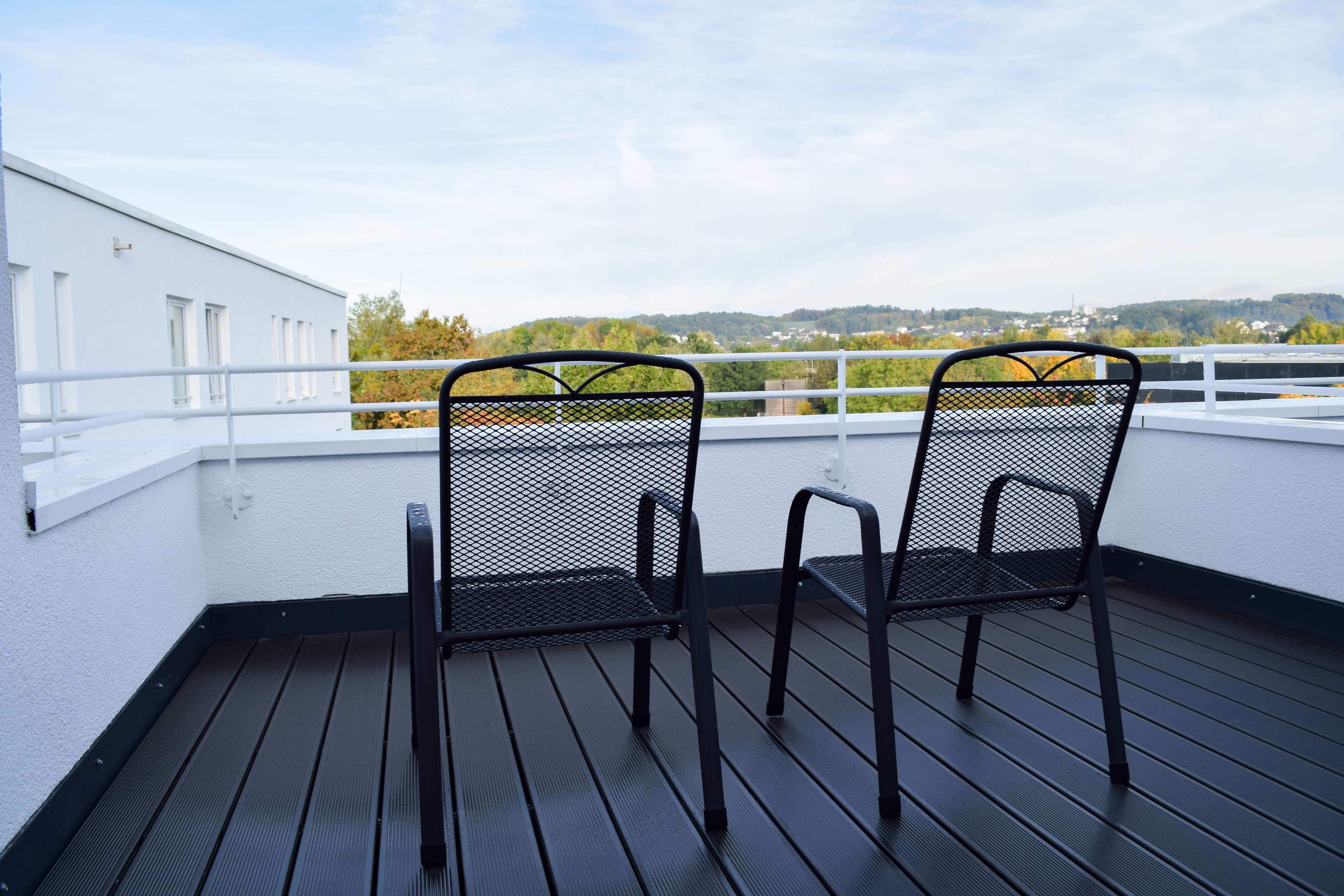 Two black metal chairs on a wooden deck with a white railing, overlooking a landscape with trees and buildings.
