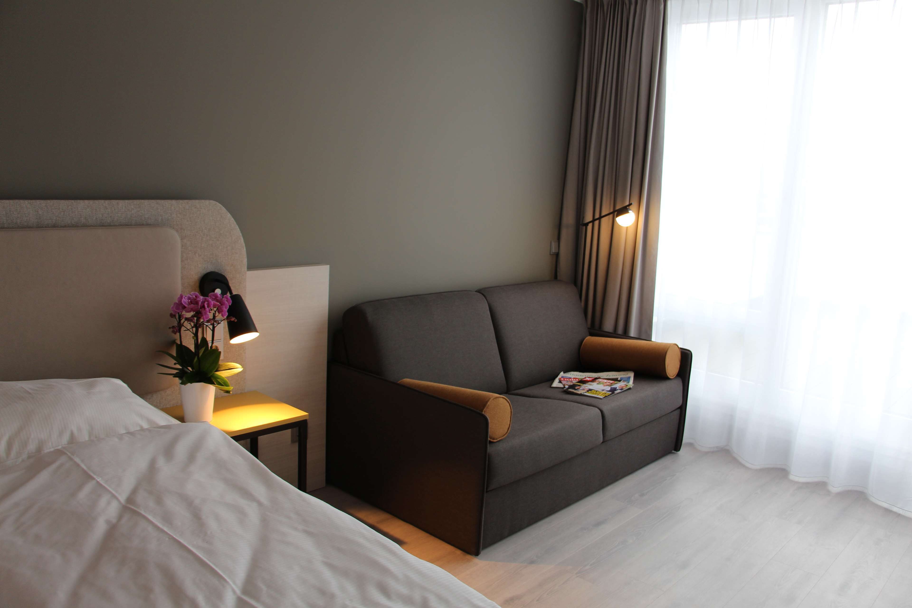 A hotel room with a bed, a sofa, a small table with a lamp, and a window with curtains.