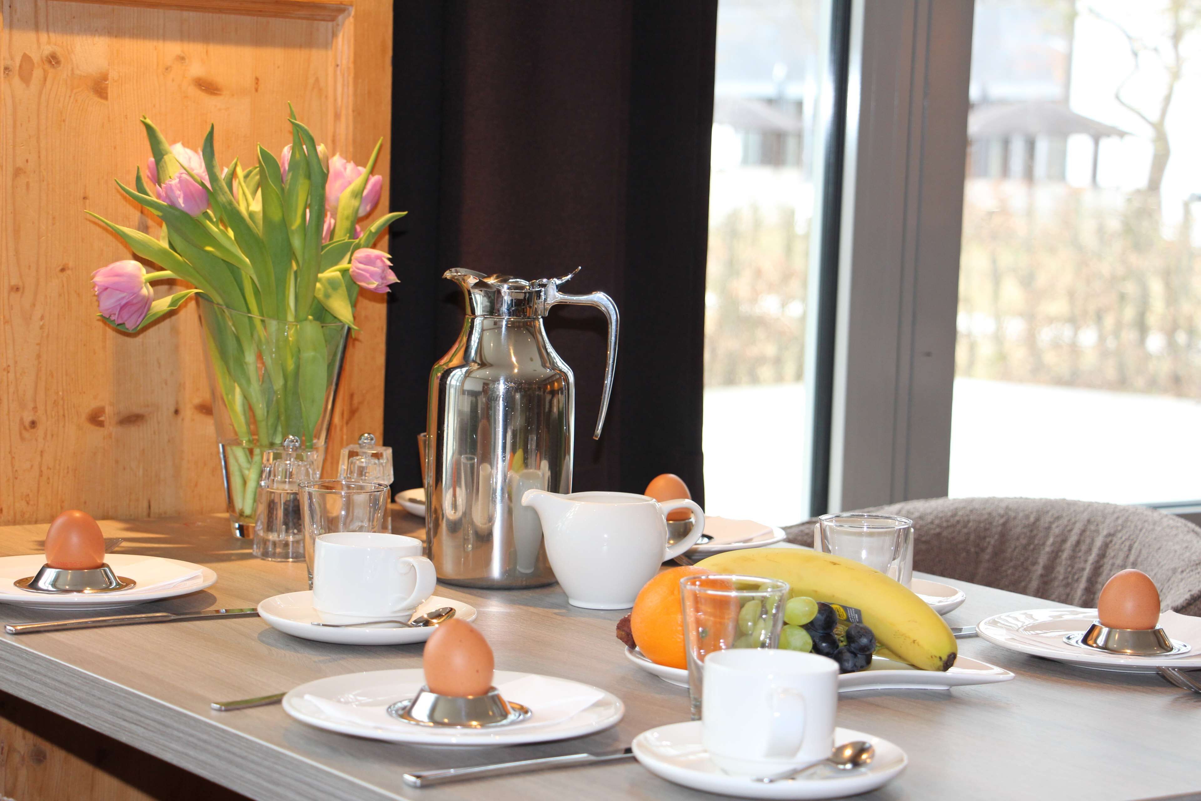 Une table de petit-déjeuner avec des assiettes, des tasses, un pichet, un vase de fleurs et une vue sur la fenêtre.