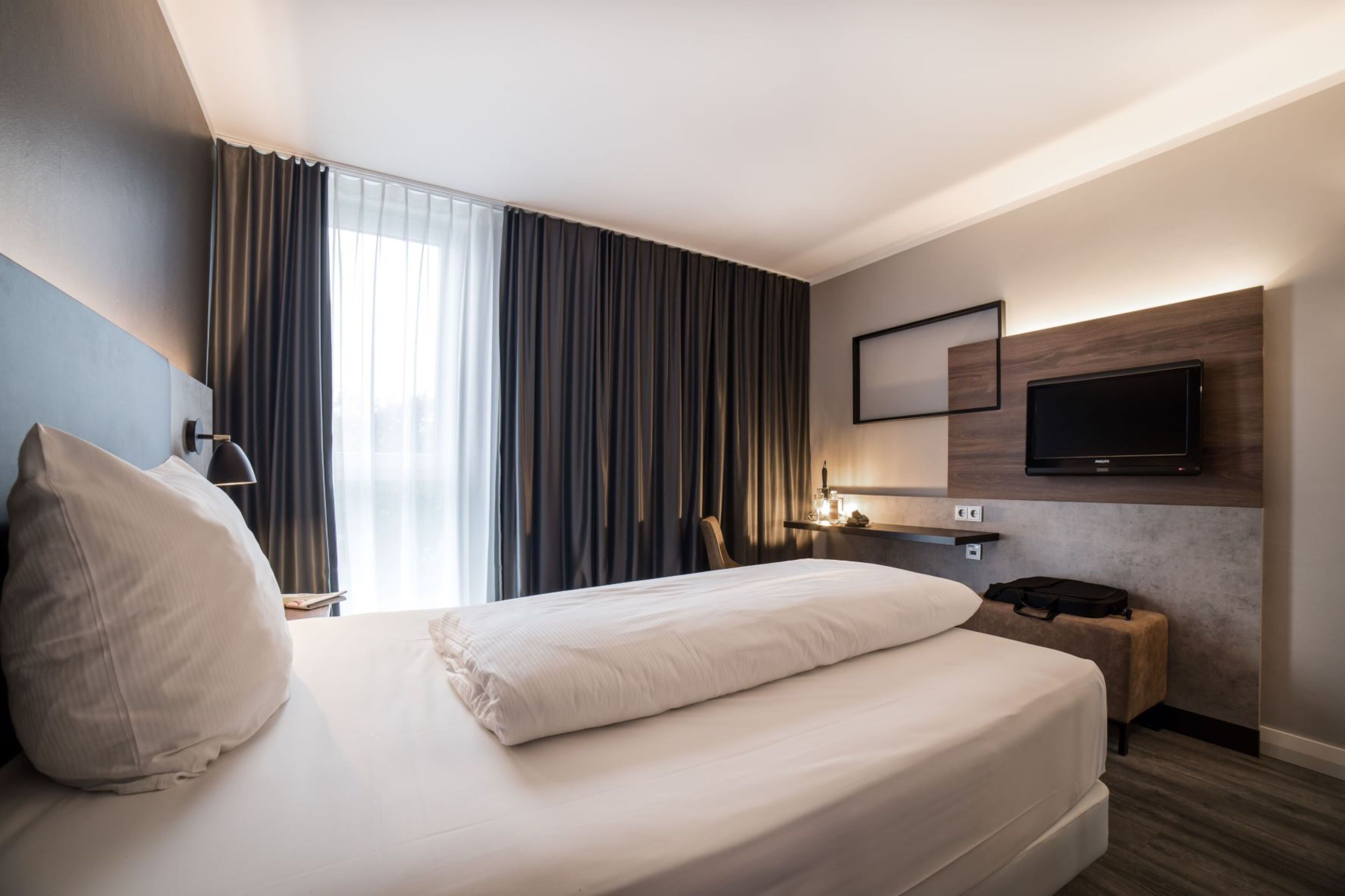 A modern hotel room with a large bed, a flat-screen TV mounted on the wall, a bedside table with a lamp, and a window with curtains.