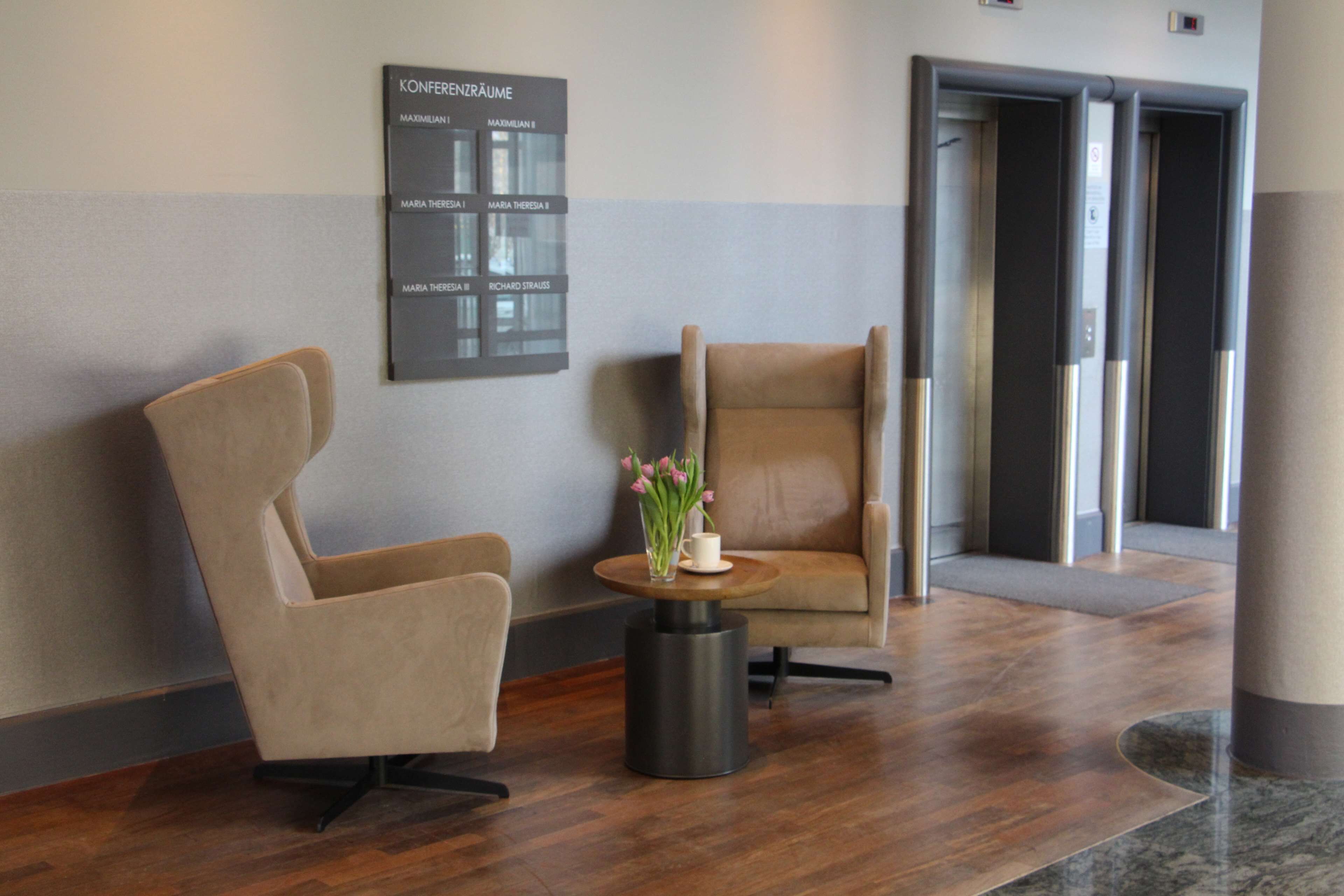 A modern lobby with two leather armchairs, a small round table with a vase of flowers, and a wall-mounted directory.