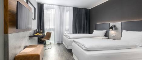 Quarto de hotel moderno com duas camas, uma escrivaninha com cadeira, um pequeno banco e uma janela grande com cortinas.