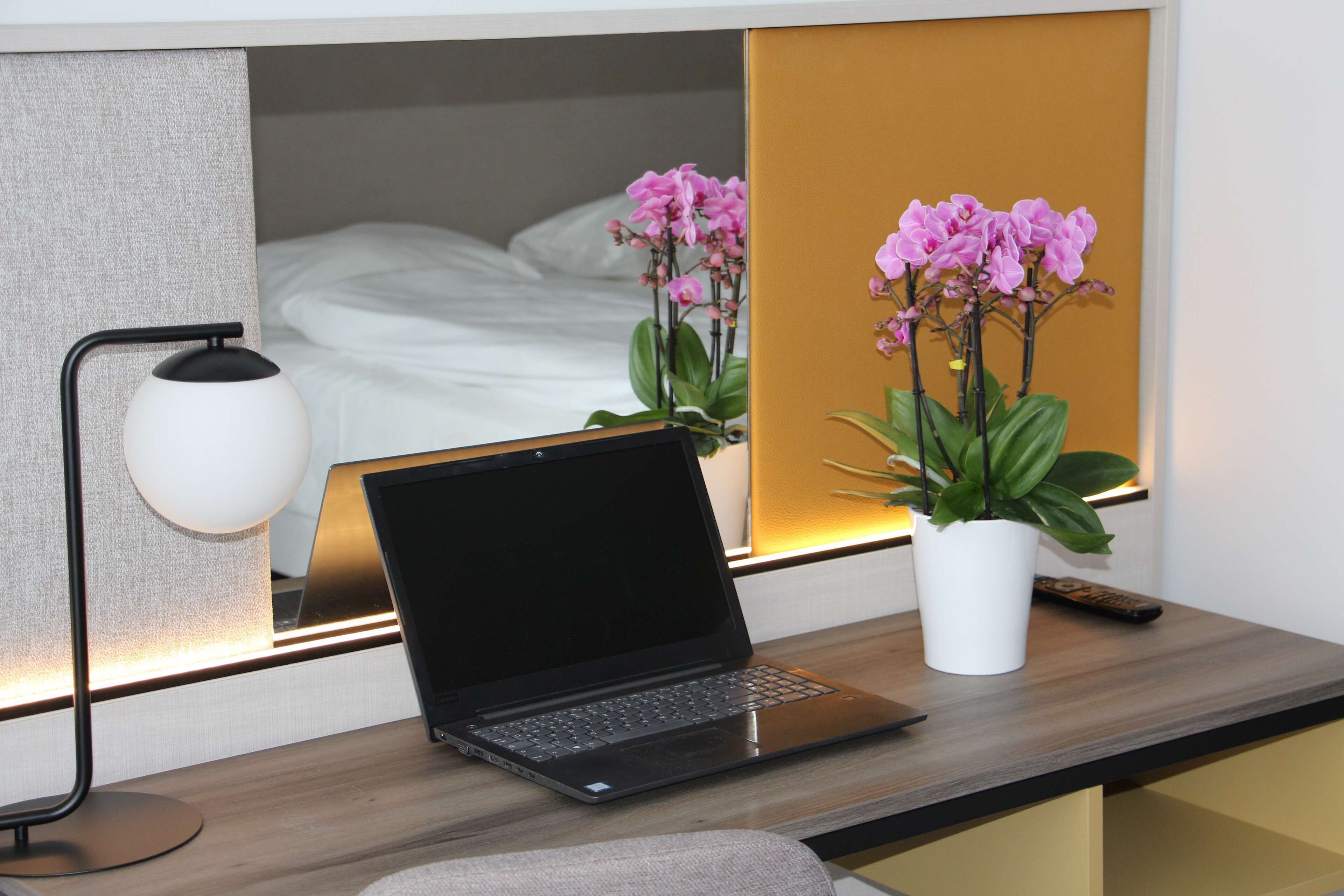Un ordinateur portable sur un bureau, avec une lampe, une plante en pot et un lit en arrière-plan.