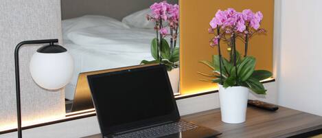 A laptop on a desk with a lamp, a potted plant, and a bed in the background.