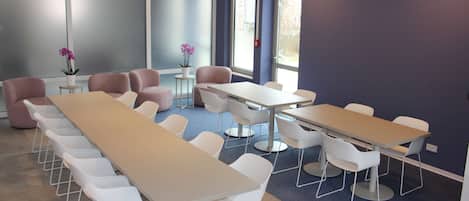 Un salón de conferencias con mesas largas y sillas blancas, ventanas amplias y una pared decorada en azul.