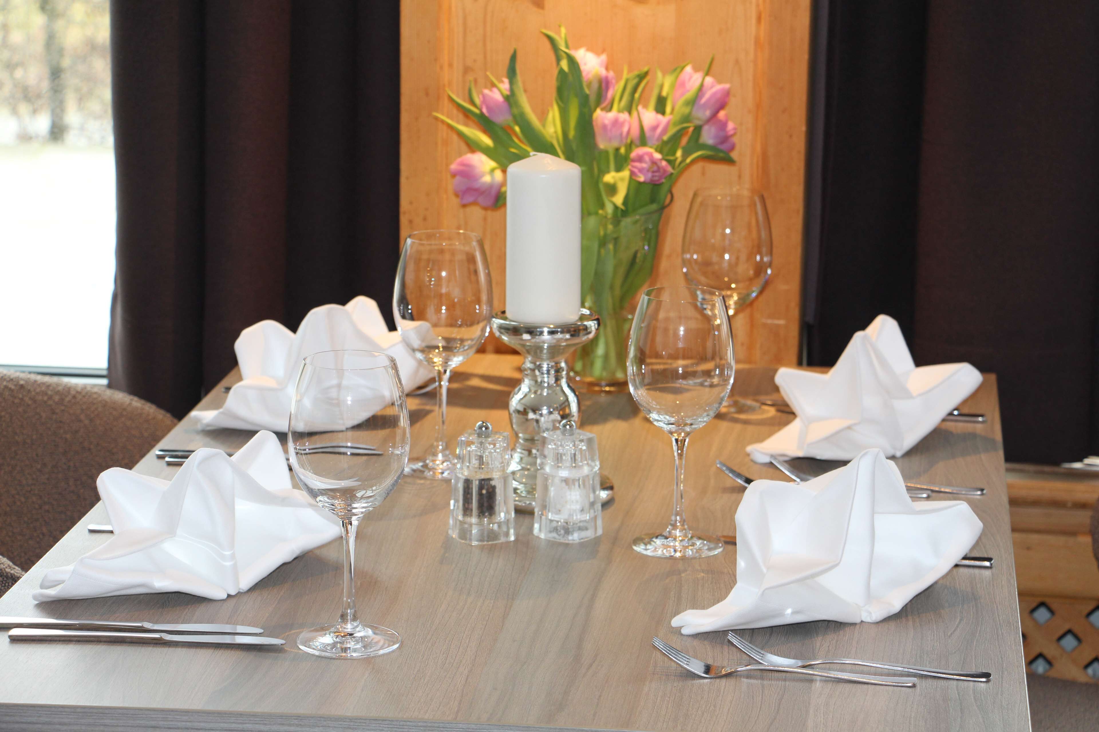Mesa de comedor dispuesta con servilletas blancas, copas de vino y cubiertos, con una vela y flores al fondo.