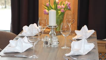 Mesa de comedor dispuesta con servilletas blancas, copas de vino y cubiertos, con una vela y flores al fondo.