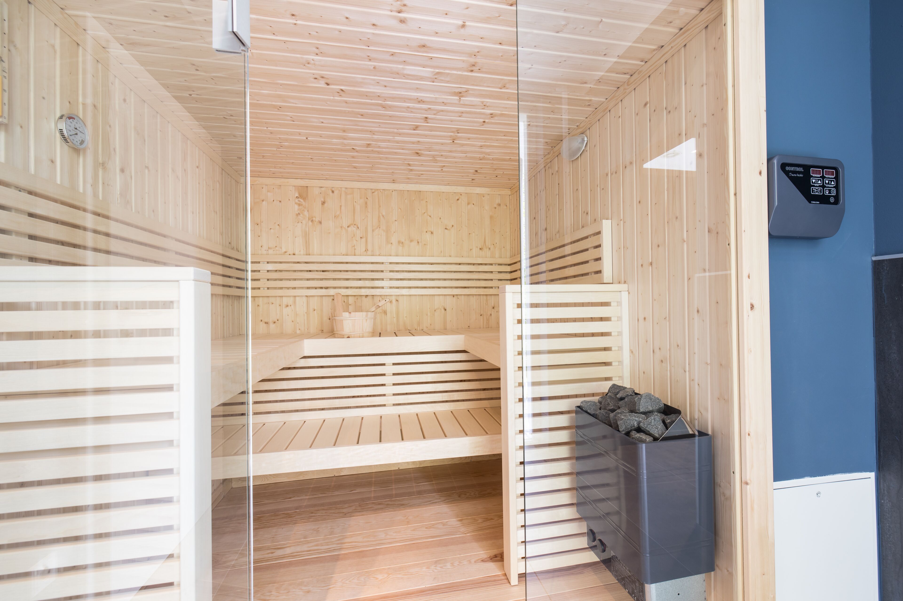 A sauna with wooden walls and a bench. There is a control panel on the wall and a trash bin with rocks.