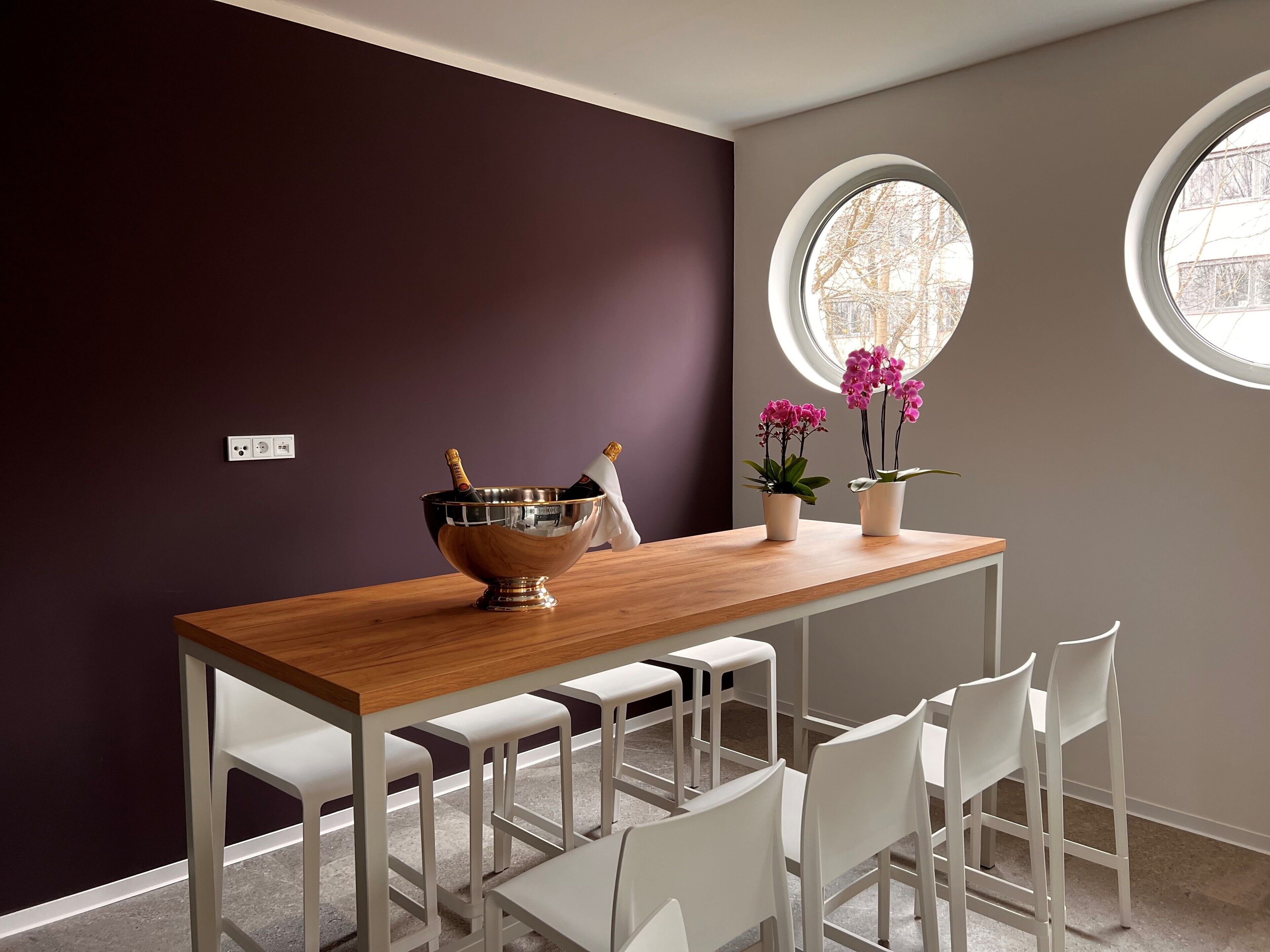 Un área de comedor con una mesa de madera, sillas blancas, un bol metálico con una botella y dos plantas en macetas.