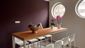 Een eethoek met een houten tafel, witte stoelen, een metalen kom met een fles en twee potplanten.
