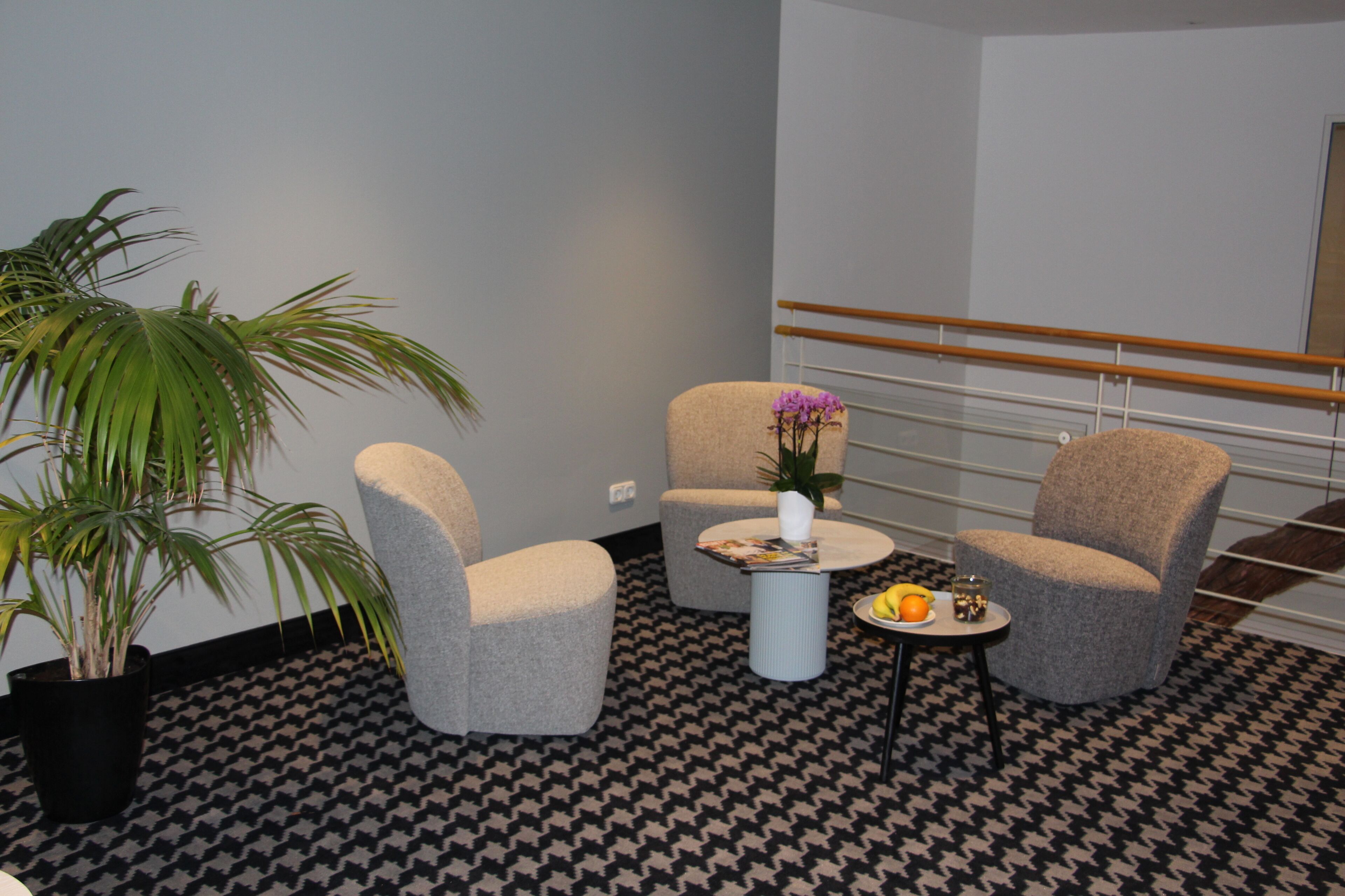 A modern hotel lobby with two grey armchairs, a coffee table with a vase and magazine, a potted plant, and a patterned carpet.