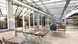 Un patio exterior con mesas y sillas, ventanas amplias y una zona cubierta.