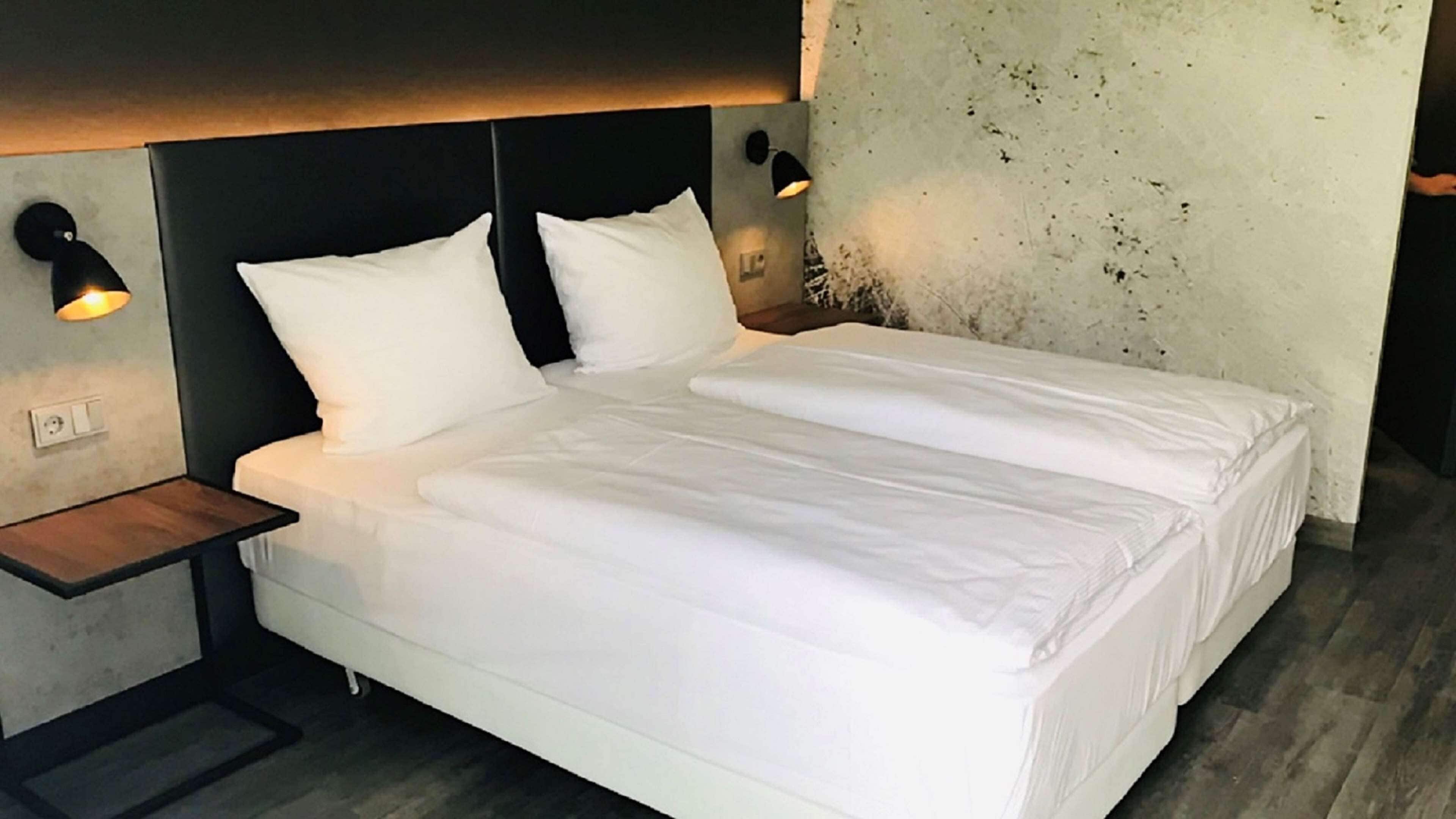 Una cama bien hecha con ropa de cama blanca en una habitación moderna con una pared de concreto y dos lámparas fijadas a la pared.