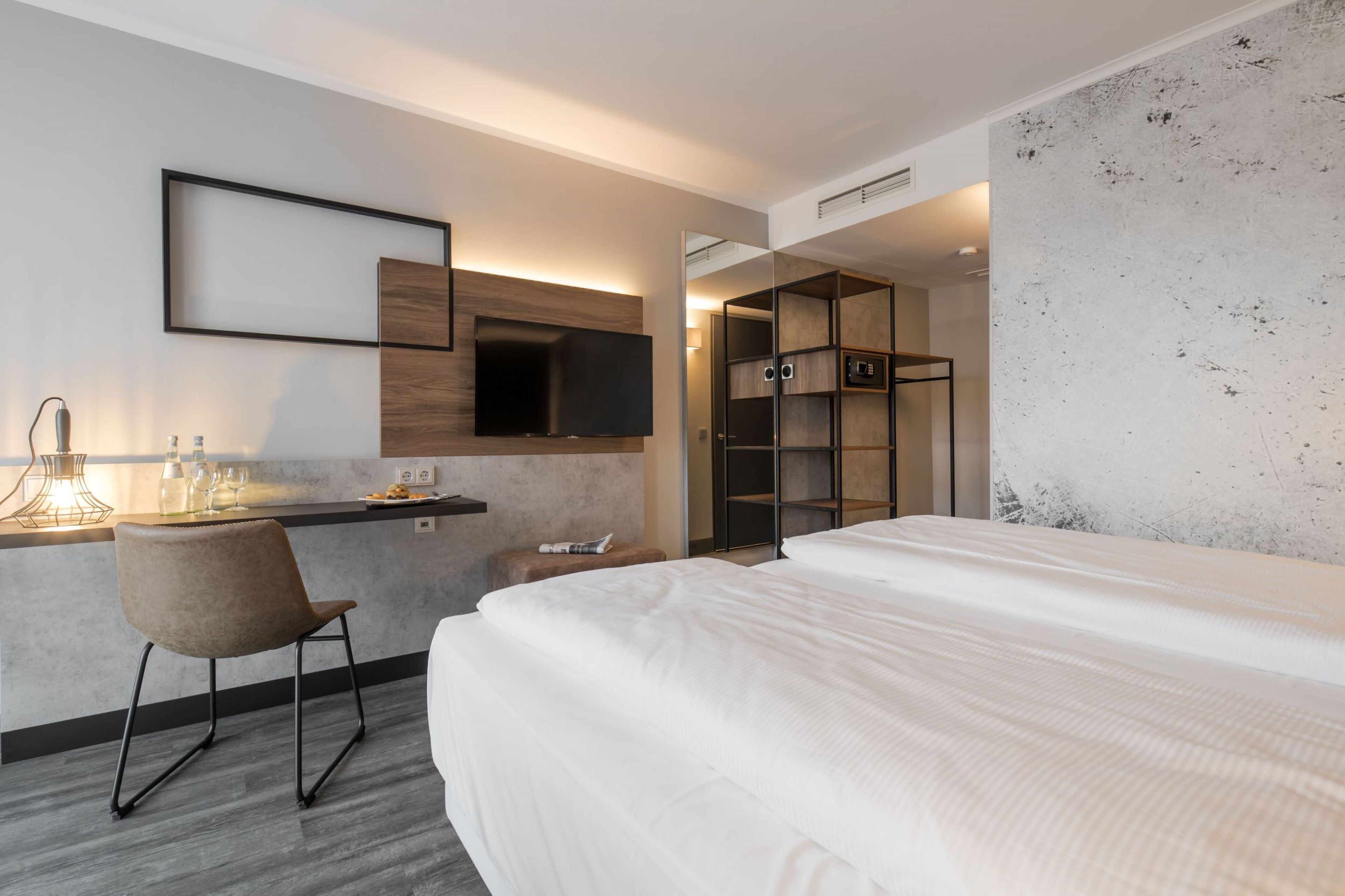 A modern hotel room with a large bed, a desk with a chair, a flat-screen TV, and built-in storage.