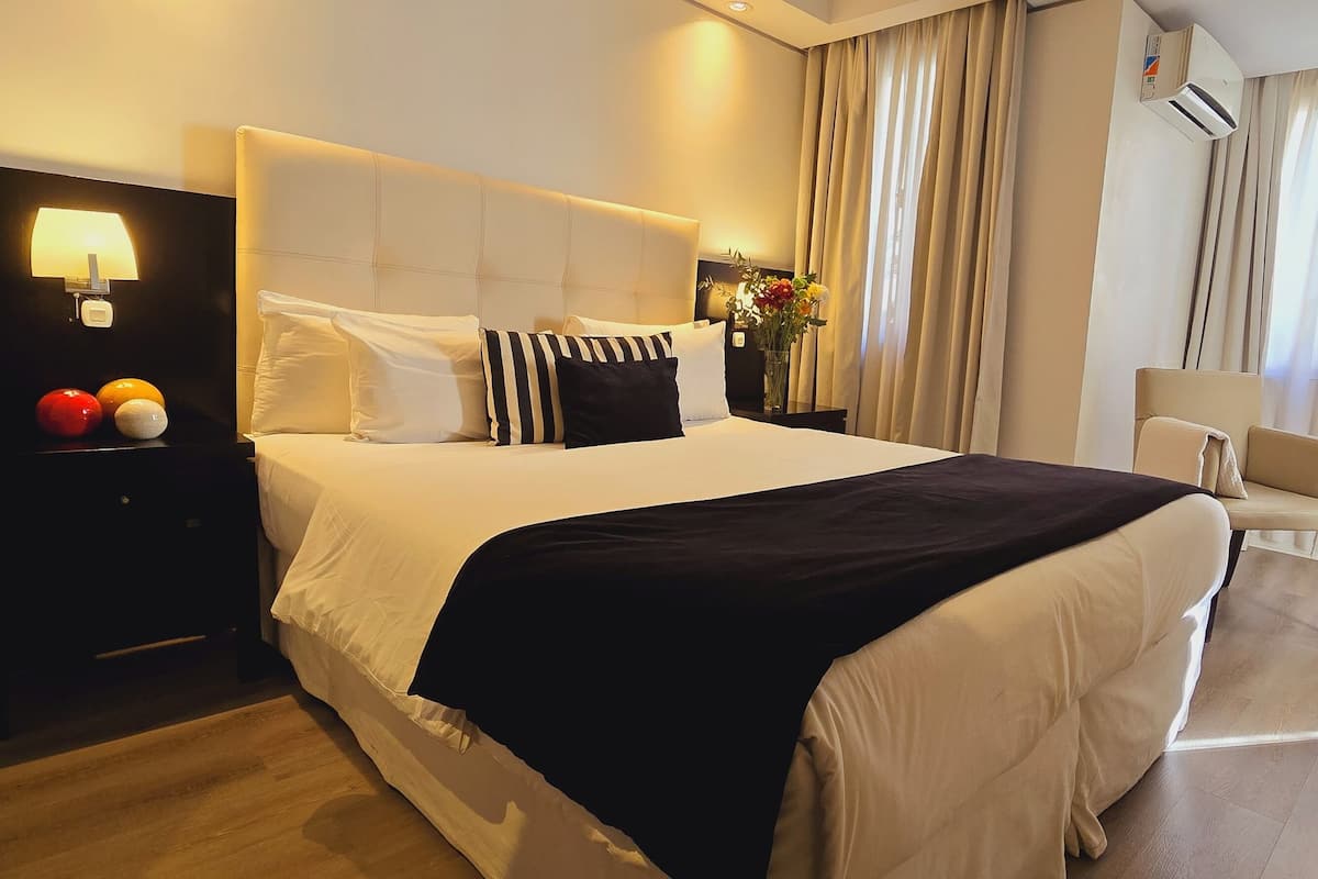 Habitación de hotel con una cama grande, una mesita de noche con lámpara, una silla y vistas al exterior a través de la ventana.