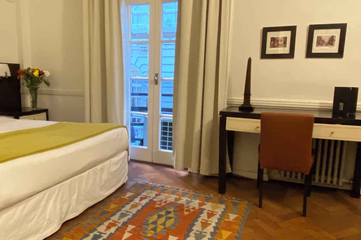 Una habitación de hotel con cama, escritorio, silla, radiador y ventana con cortinas.