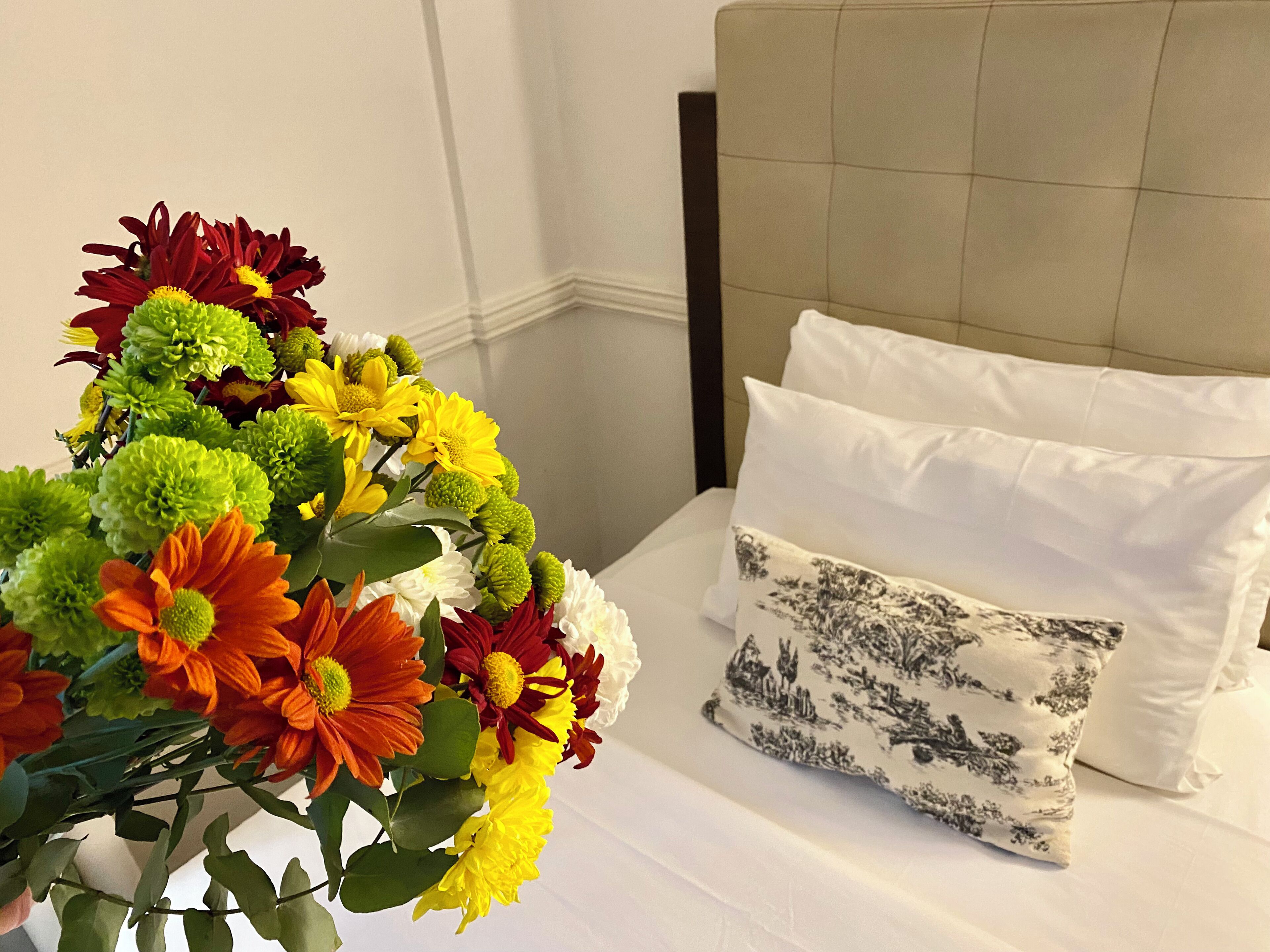 Una cama con ropa de cama blanca y un cojín floral, un ramo de flores en primer plano.