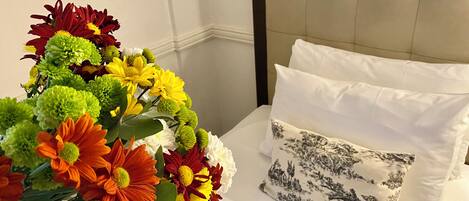 Una cama con ropa de cama blanca y un cojín floral, un ramo de flores en primer plano.