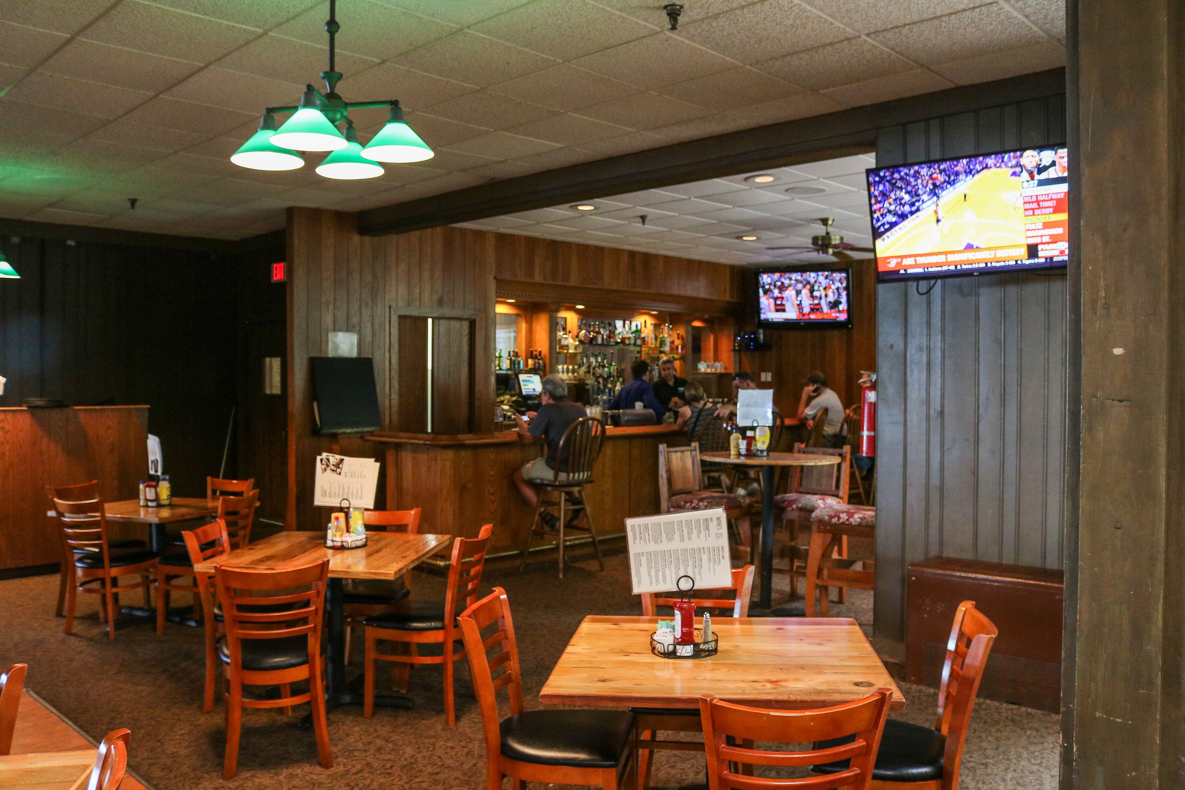 A restaurant with wooden tables and chairs, a bar area, and a television displaying a sports game.