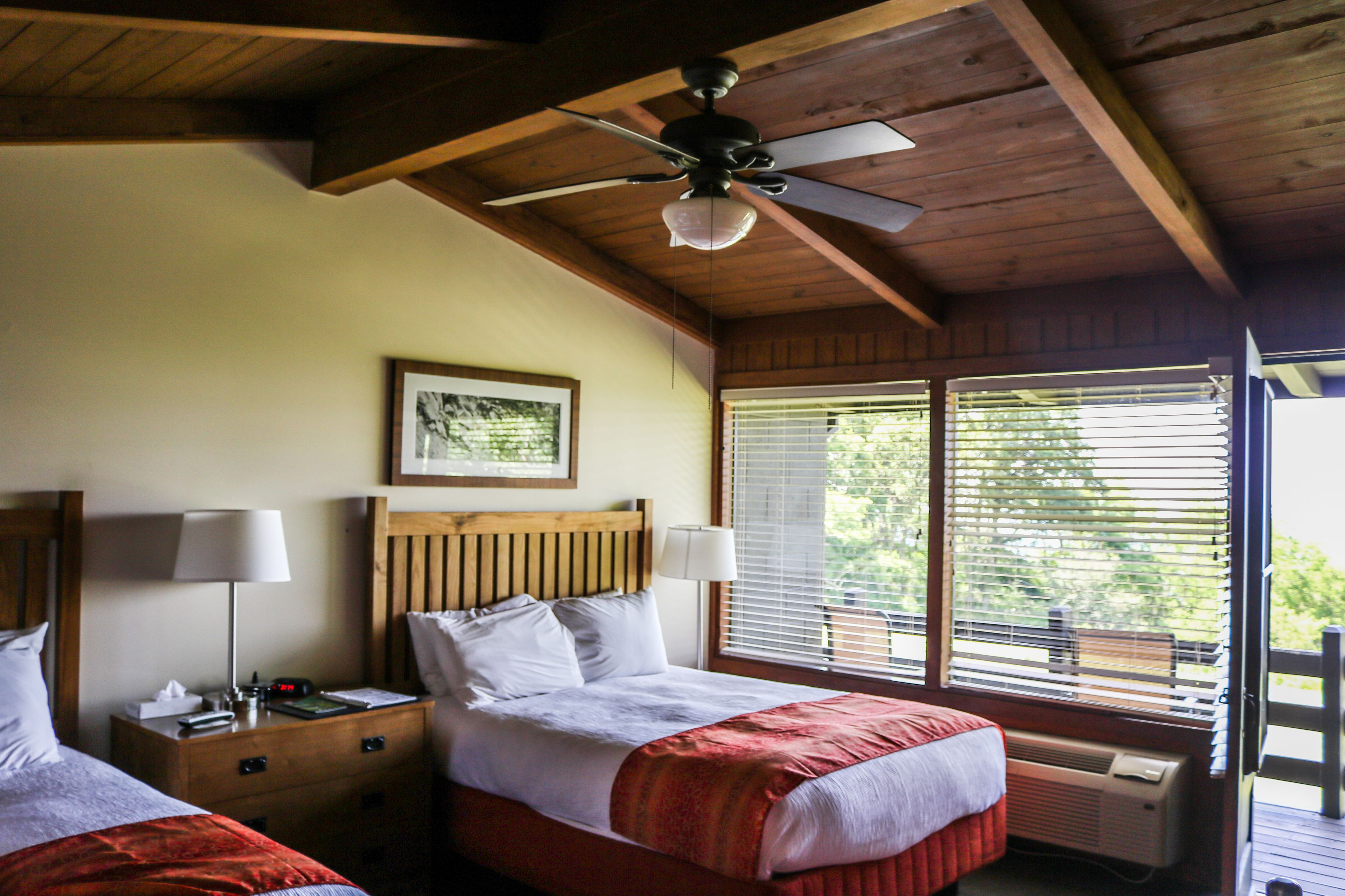 A bedroom with two beds, wooden headboards, a ceiling fan, a framed picture on the wall, and a window with blinds.