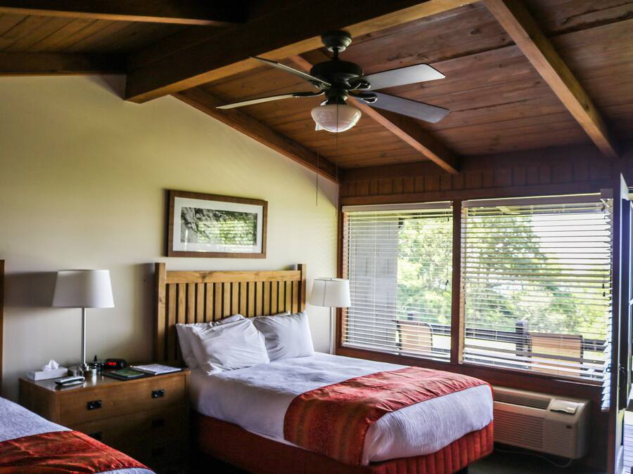 A bedroom with two beds, wooden headboards, a ceiling fan, a framed picture on the wall, and a window with blinds.