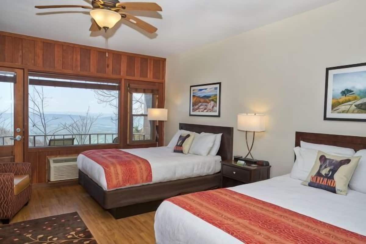 A room with two beds, a ceiling fan, a window with a view, and a framed picture on the wall.