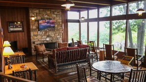 A cozy interior with a stone fireplace, wooden furniture, and large windows offering a view of the outdoors. - Skyland (Luray)
